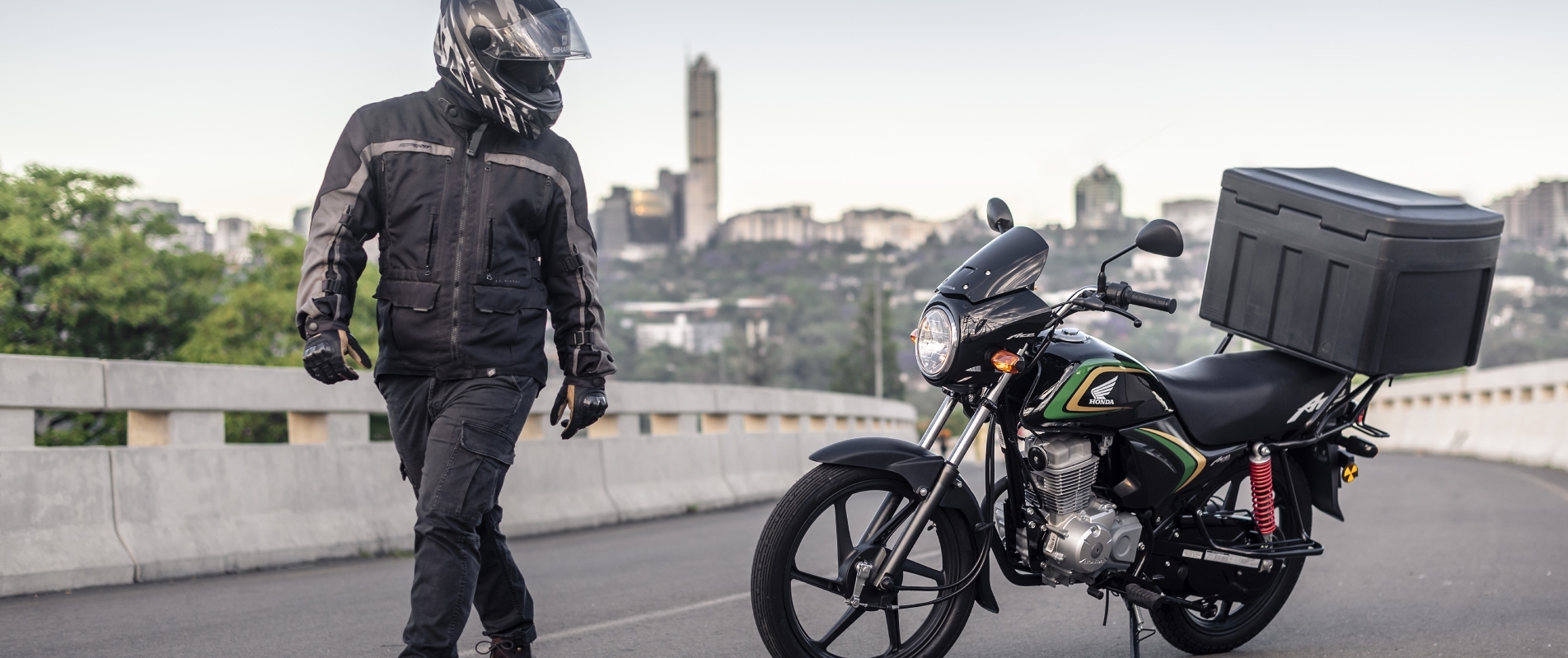 Rider walking toward a Honda Ace with delivery box on a Johannesburg overpass